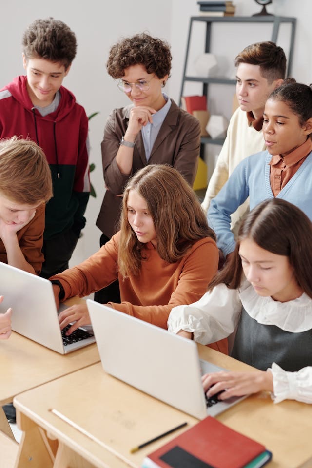 Abbildung zeigt Kinder die in der Klasse am Computer arbeiten. Andere Schüler*innen schauen auf den Bildschirm.