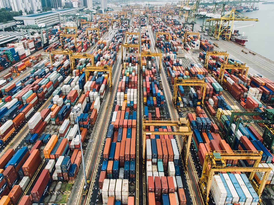 Ein großer Hafen mit einer großen Menge an bunten Containern, Stapelweise aufgetürmt, rechts das Meer mit Frachtern, die sich für den Transport bereit halten.