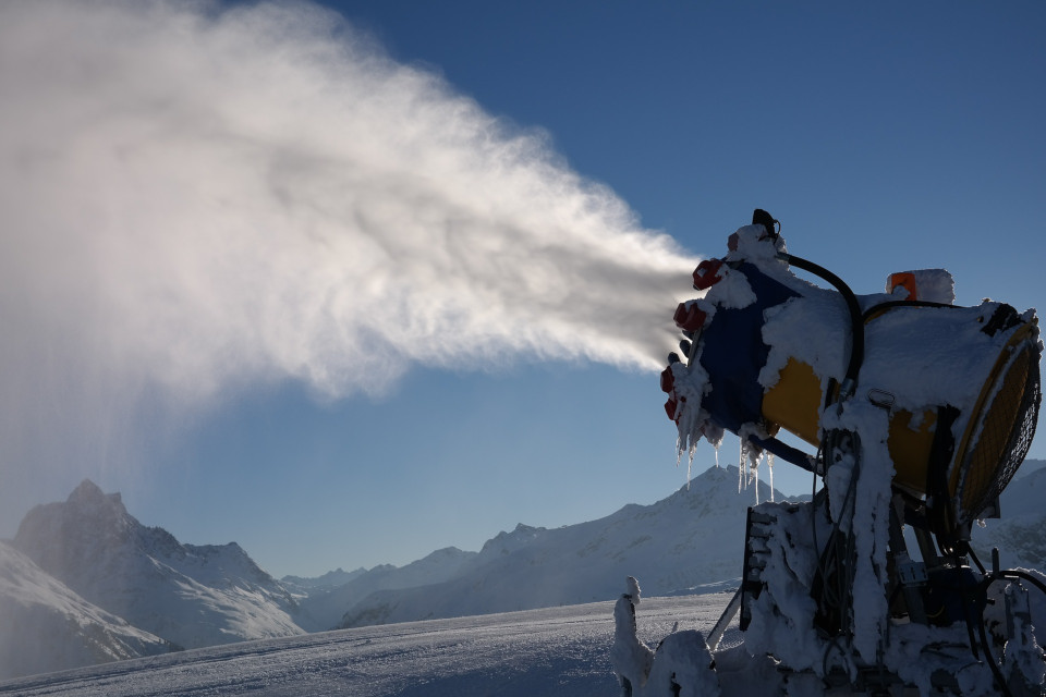 Abbild einer vereisten Schneekanone, welche künstlichen
   Schnee verteilt und eine kahle Gebirgslandschaft im Huntergrund.